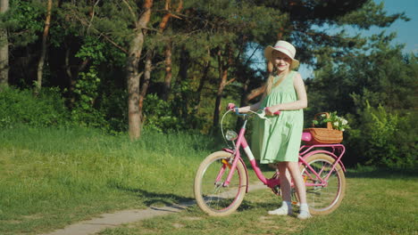 A-Girl-In-A-Summer-Dress-Is-Standing-Near-A-Bicycle-Looking-At-The-Camera-With-Her-Basket-Of-Wildflo