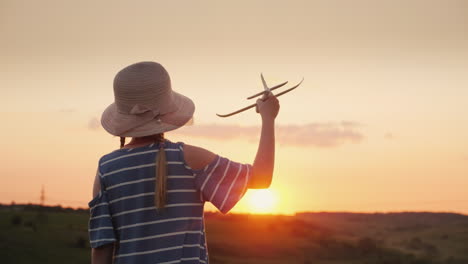 Ein-Mädchen-Mit-Zöpfen-Und-Einem-Hut,-Das-Bei-Sonnenuntergang-Mit-Einem-Holzflugzeug-Spielt-Der-Traum-Von-Fernreisen