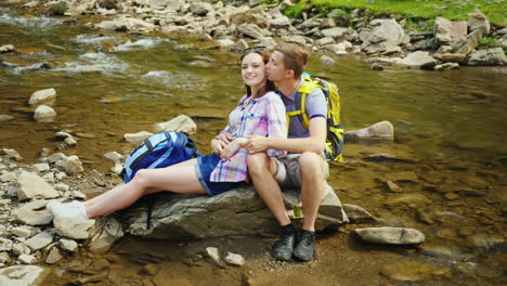 A-Loving-Couple-Of-Tourists-With-Backpacks-Rests-In-A-Picturesque-Place-Near-A-Mountain-River-They-E