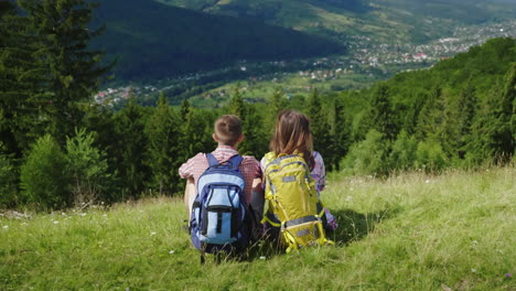 Young-Couple-Of-Tourists-With-Backpacks-Sitting-On-A-Green-Meadow-Looking-At-A-Picturesque-Mountain-