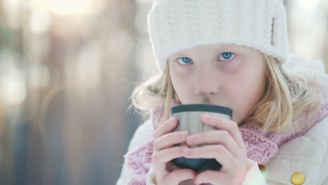 A-Little-Girl-In-A-Warm-Knitted-Hat-Drinks-Hot-Tea-From-A-Thermos-In-The-Winter-Forest-Portrait-In-T