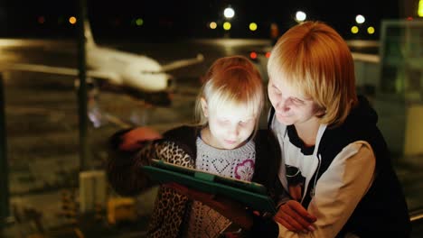 Mom-And-Daughter-Play-On-The-Tablet-At-The-Airport-Late-Evening-The-Airliner-Is-Visible-Outside-The-