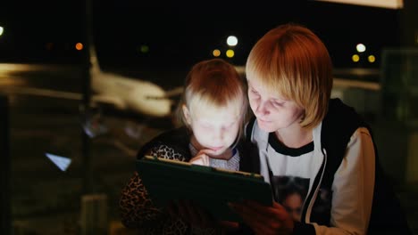 Mom-And-Daughter-Play-On-The-Tablet-At-The-Airport-Late-Evening-The-Airliner-Is-Visible-Outside-The-