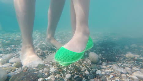 Legs-In-Protective-Shoes-On-The-Pebble-Bottom-Of-The-Sea-Safe-Bathing-In-The-Sea-Concept