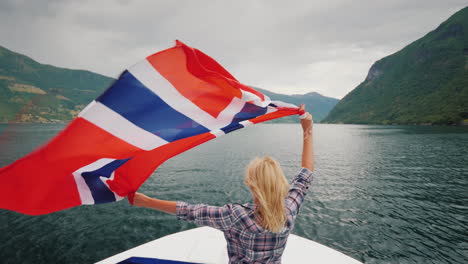 An-Active-Woman-With-A-Large-Norwegian-Flag-Is-Standing-On-The-Bow-Of-A-Cruise-Ship-Fjord-Cruise-In-