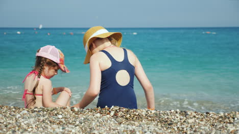 Two-Girls-Are-Sitting-On-A-Pebble-By-The-Sea-Looking-For-Beautiful-Pebbles-Holidays-With-Children-At
