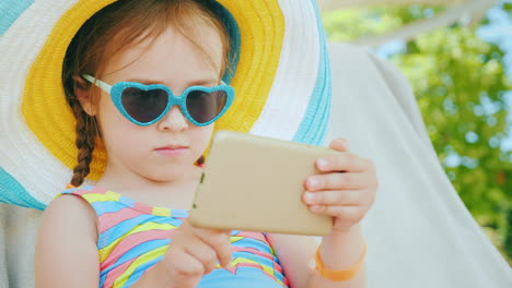 On-The-Beach-With-A-Smartphone-Little-Girl-Is-Playing-On-Smartphone-Is-Sitting-In-Big-Hat-In-Chaise-
