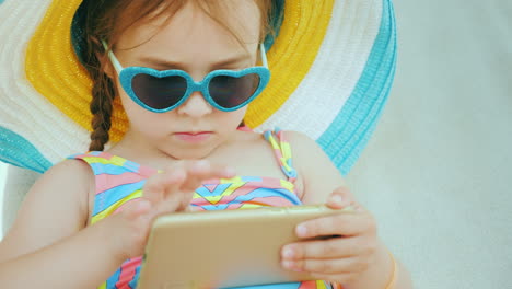 On-The-Beach-With-A-Smartphone-Little-Girl-Is-Playing-On-Smartphone-Is-Sitting-In-Big-Hat-In-Chaise-