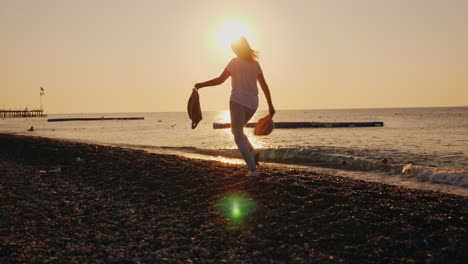 Carefree-Woman-With-A-Backpack-And-Hat-In-Her-Hands-Runs-Along-The-Seashore-At-Dawn-Summer-And-Vacat