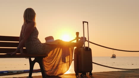 A-Romantic-Woman-In-A-Light-Dress-With-A-Burly-Bag-Sits-On-The-Dock-Looks-At-The-Sunrise-Over-The-Se