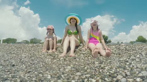 A-Young-Mother-In-A-Cool-Colorful-Hat-With-Two-Children-Sitting-On-The-Coast-Playing-Fun-With-The-Su