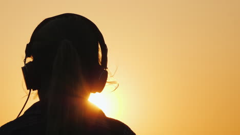 Silhouette-Of-A-Girl-In-Headphones-Listening-To-Music-Against-The-Backdrop-Of-A-Large-Setting-Sun-An