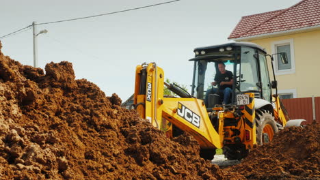 Excavator-Digs-A-Hole-For-Laying-Communications-Against-The-Background-Of-A-Modern-Two-Story-House