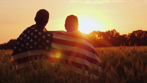 A-Man-And-His-Son-Admire-The-Sunset-Over-A-Field-Of-Wheat-Wrapped-In-The-Flag-Of-The-Usa
