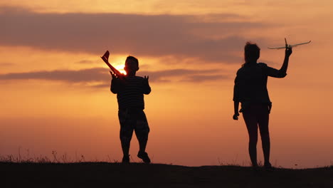 Silhouettes-Of-A-Girl-And-A-Boy-Playing-Together-With-Airplanes-At-Sunset-A-Happy-And-Carefree-Niño