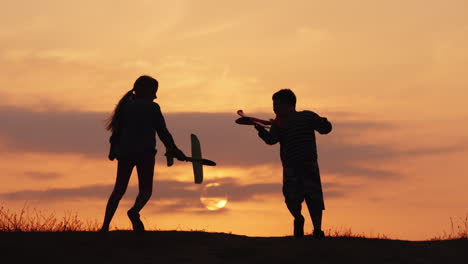 Silhouetten-Eines-Mädchens-Und-Eines-Jungen,-Die-Bei-Sonnenuntergang-Mit-Flugzeugen-Zusammen-Spielen,-Ein-Glückliches-Und-Sorgloses-Kind