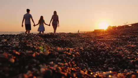 Young-Family-With-A-Child-Walking-Along-The-Beach-At-Sunset-In-The-Foreground-A-Pebble-Beach