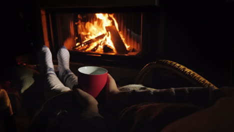A-Man-Warms-His-Feet-By-The-Fireplace-Holds-In-His-Hands-A-Red-Cup-With-A-Hot-Drink