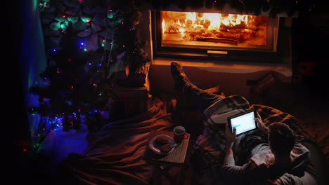 Top-View-A-Man-Uses-A-Tablet-By-The-Fireplace-And-Christmas-Tree-Ordering-Holiday-Gifts