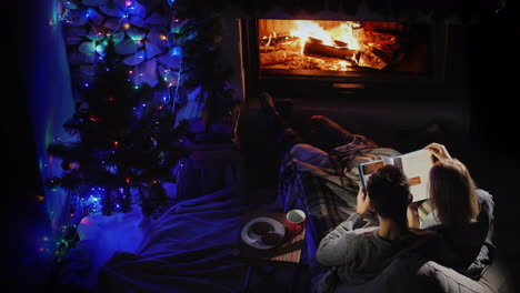 A-Man-And-A-Woman-Read-A-Book-By-The-Fireplace-Where-There-Is-A-Christmas-Tree
