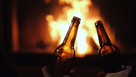 Zwei-Flaschen-Bier-In-Den-Händen-Von-Männern-Auf-Einem-Feuerhintergrund-In-Einer-Kamin-Nahaufnahme