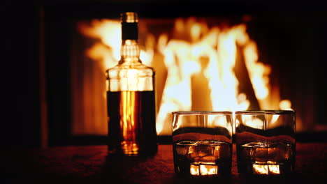 Two-Glasses-And-A-Bottle-Of-Whiskey-Stand-On-The-Table-Against-The-Background-Of-A-Hot-Fireplace