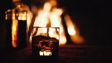 Ein-Glas-Whisky-und-Eine-Flasche-Auf-Dem-Tisch-Im-Hintergrund-Ein-Feuer-Brennt-Im-Kamin