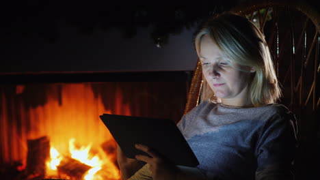 A-Young-Woman-Uses-A-Digital-Tablet-In-A-Cozy-House-Near-The-Fireplace-Where-The-Flame-Is-Burning
