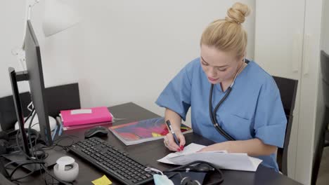 Young-beautiful-blond-female-doctor-in-blue-coat--working-at-desk-using-computer-and-doing-paperwork