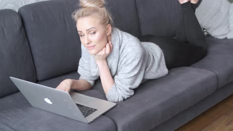 Happy-blond-woman-lying-prone-on-sofa-and-working-on-laptop-computer