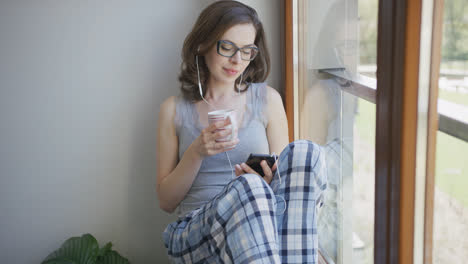 Young-woman-sitting-by-window-using-smartphone-with-earphones