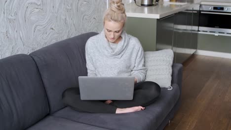 Cheerful-young-blond-woman-sitting-on-couch-in-living-room-and-using-laptop